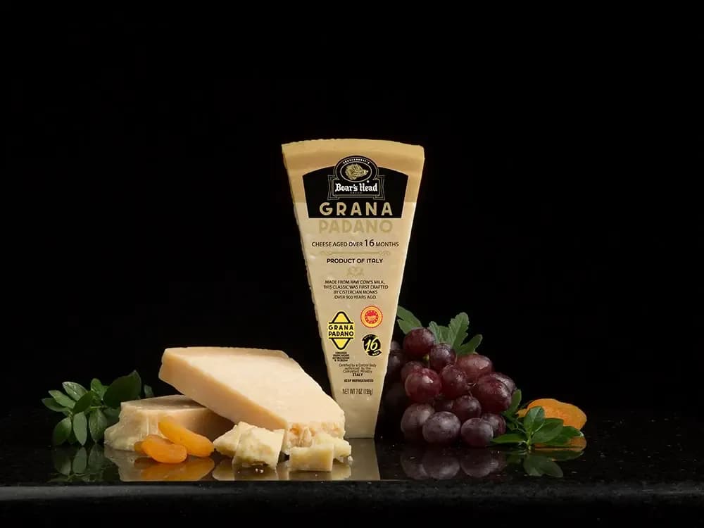 The Boar's Head® Grana Padano Cheese sits on a counter top, surrounded by grapes, dried apricots, and some unwrapped and crumbled product, with a black background.