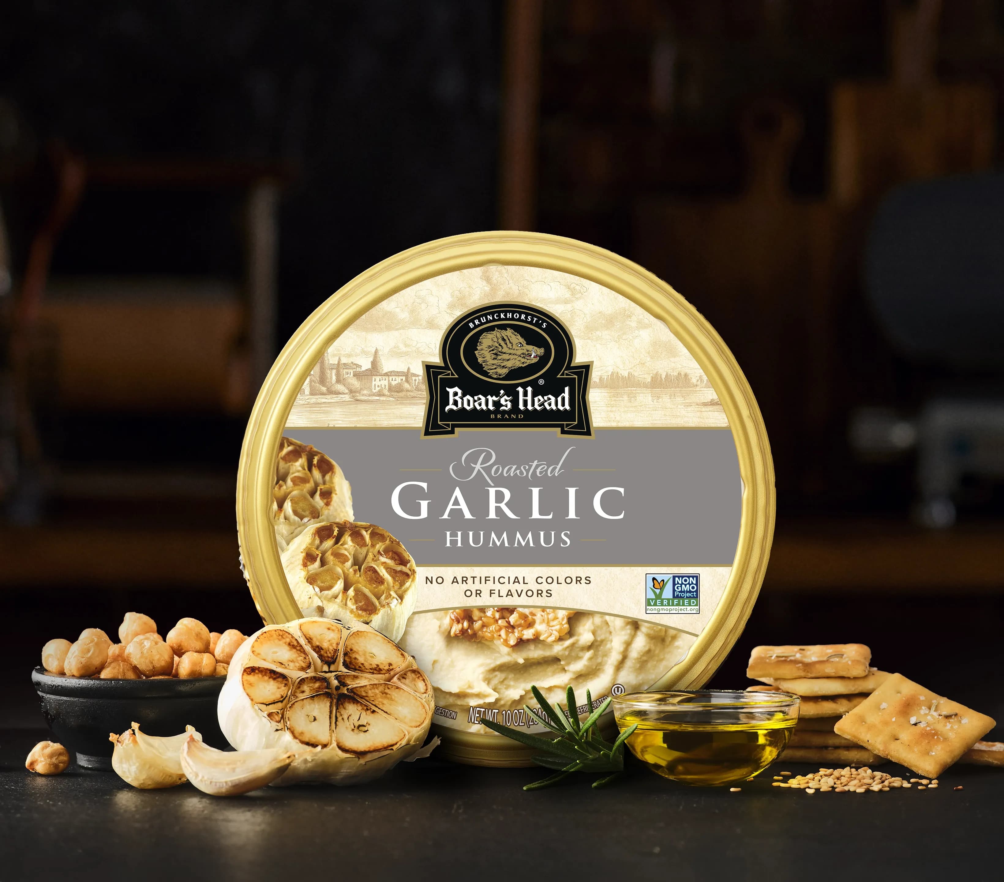 The Boar's Head® Garlic Hummus sits on a counter top, surrounded by garlic cloves, chickpeas, chips, and olive oil, all in front of blurred deli scene in the background.