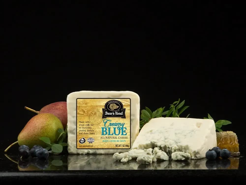 The Boar's Head® Creamy Blue Cheese sits on a counter top, surrounded by pears, blueberries, greenery, and crumbled product, all in front of black background.