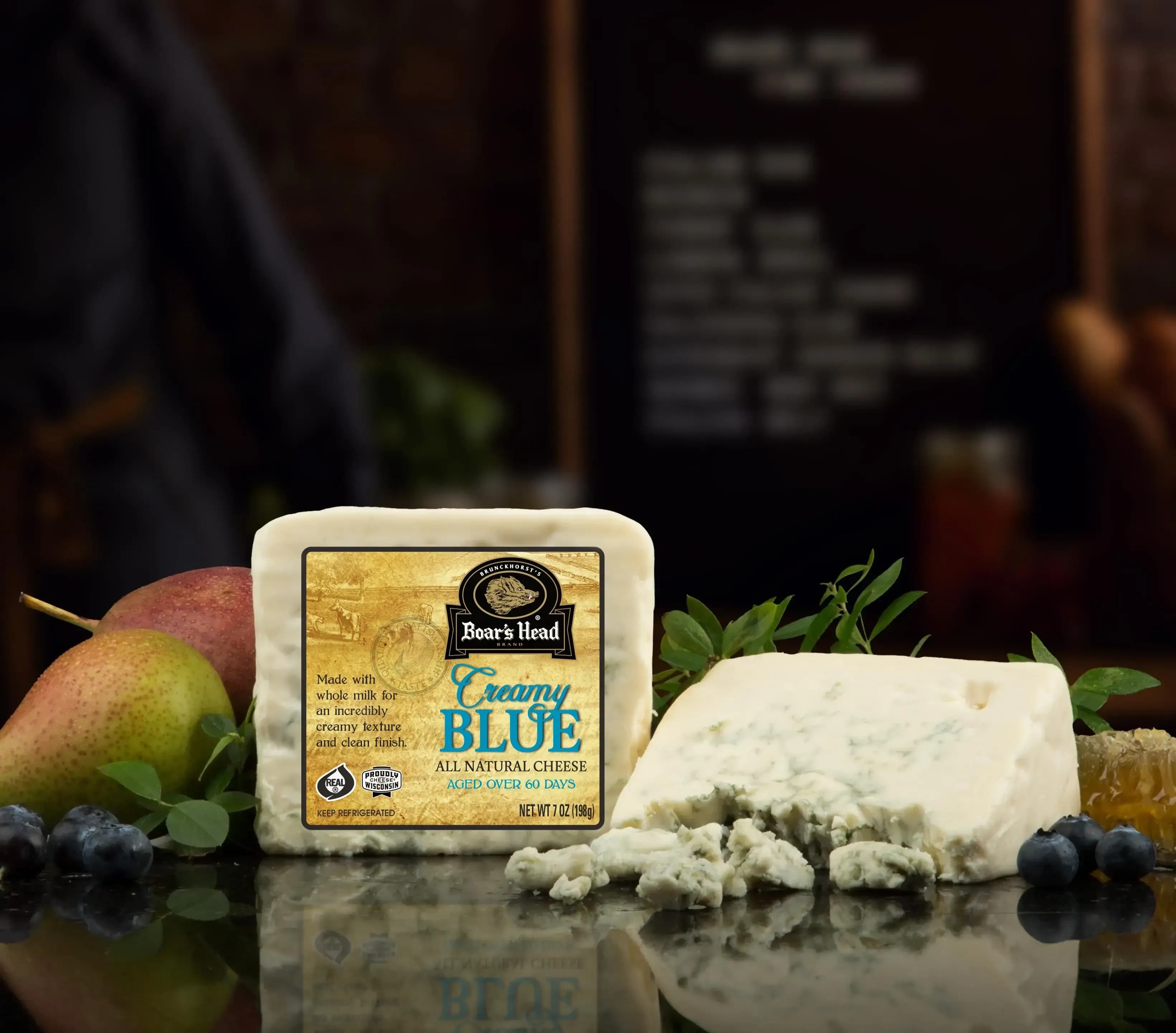 The Boar's Head® Creamy Blue Cheese sits on a counter top, surrounded by pears, blueberries, greenery, and crumbled product, all in front of blurred deli scene in the background.