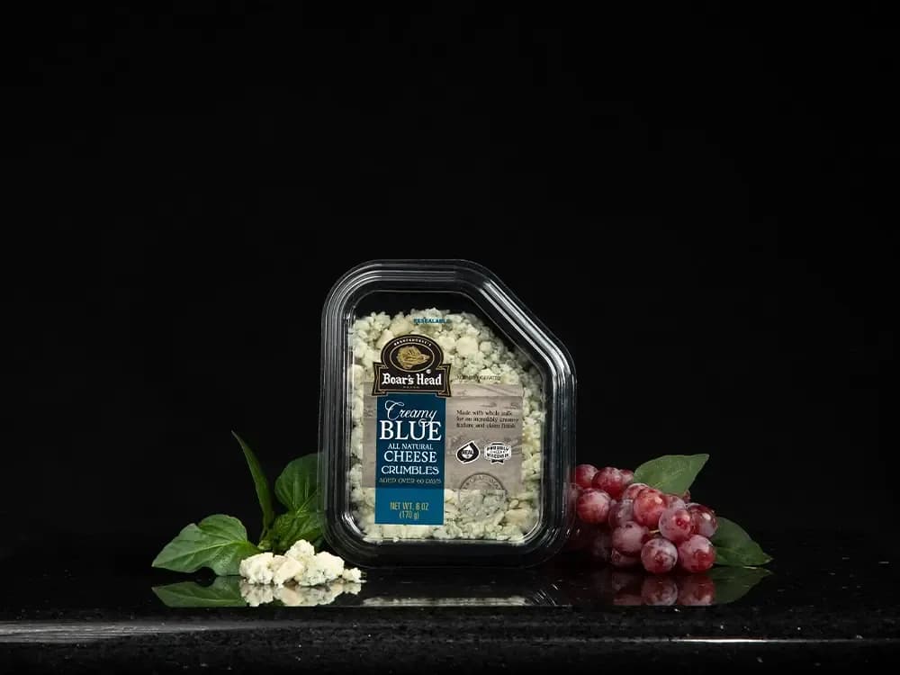 The Boar's Head® Creamy Blue Cheese Crumbles sits on a counter top, surrounded by grapes,, greenery, and crumbled product, all in front of black background.