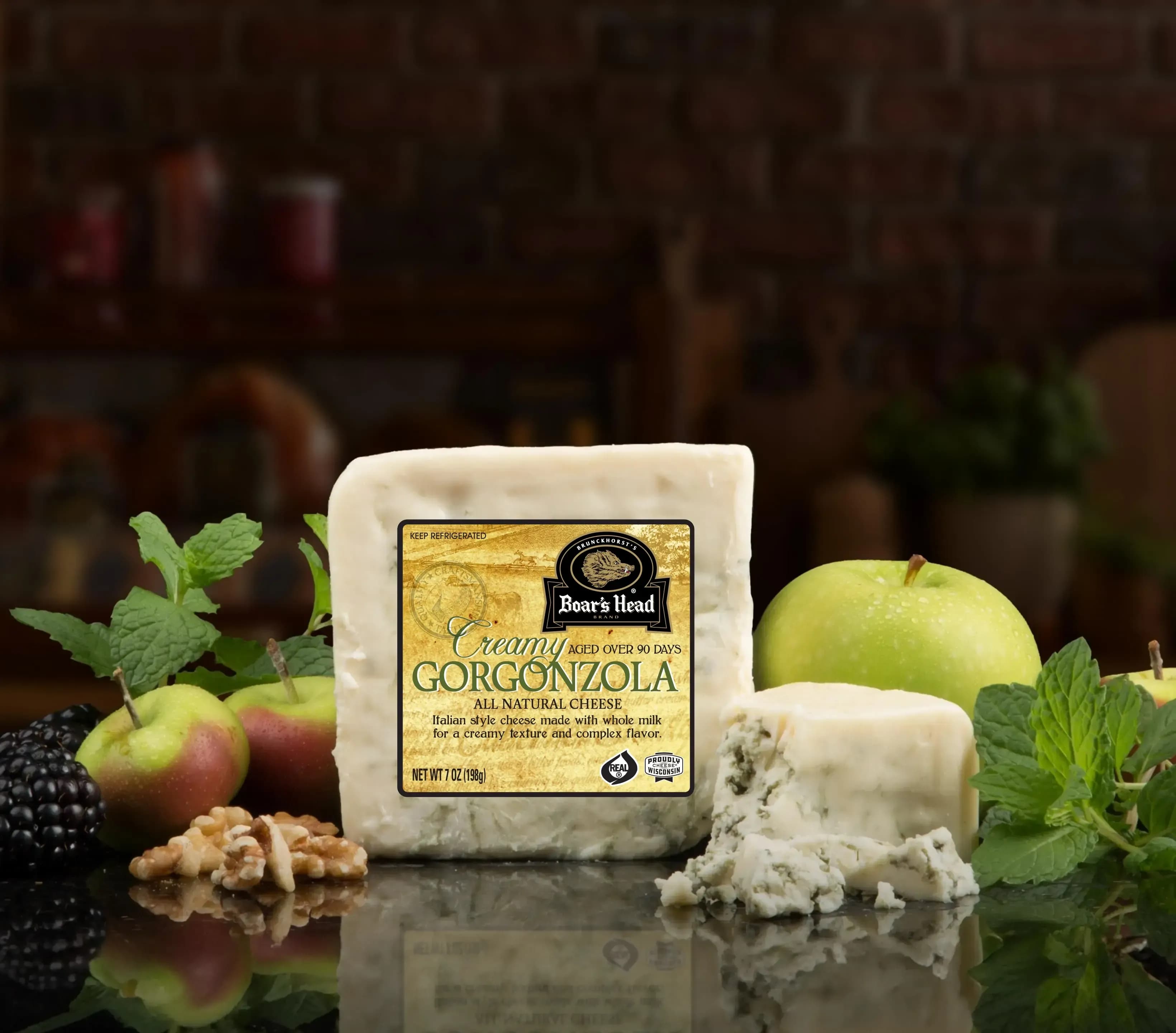 The Boar's Head® Creamy Gorgonzola Cheese sits on a counter top, surrounded by green apples, pears, nuts, greenery, and crumbled product, all in front of blurred deli scene in the background.