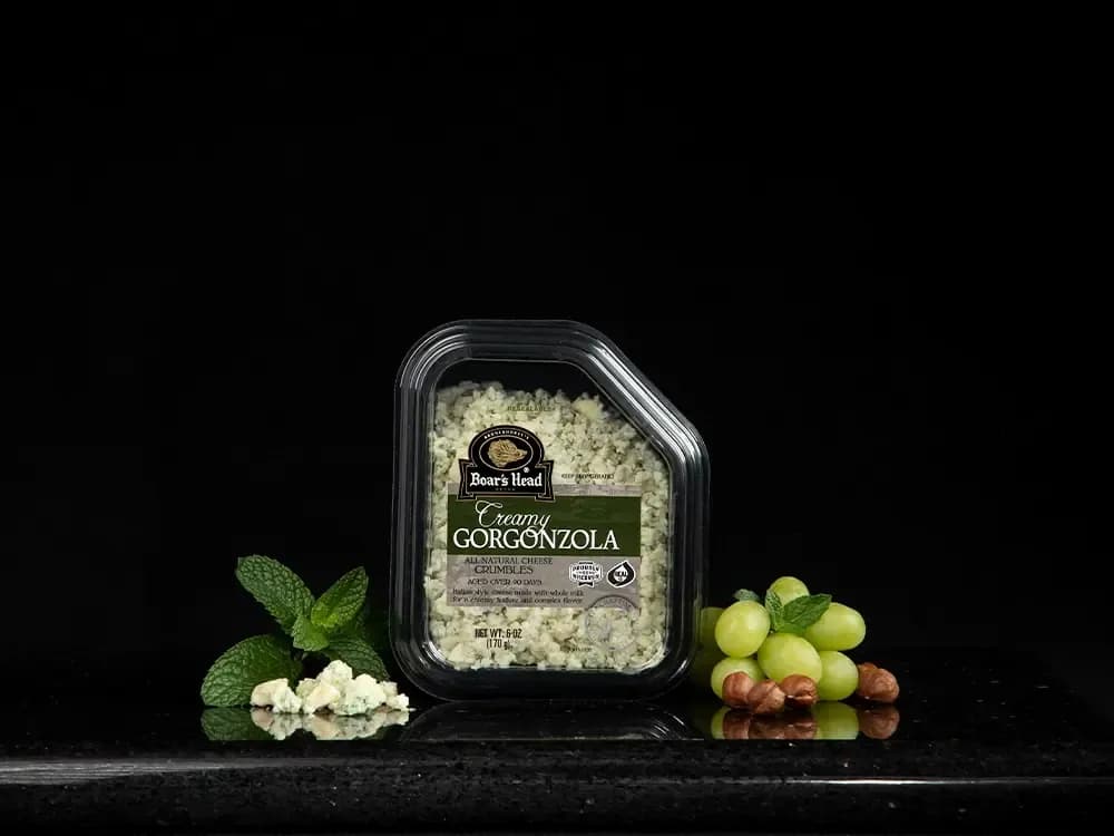 The Boar's Head® Creamy Gorgonzola Cheese Crumbles sits on a counter top, surrounded by grapes, nuts, greenery, and crumbled product, all in front of black background.