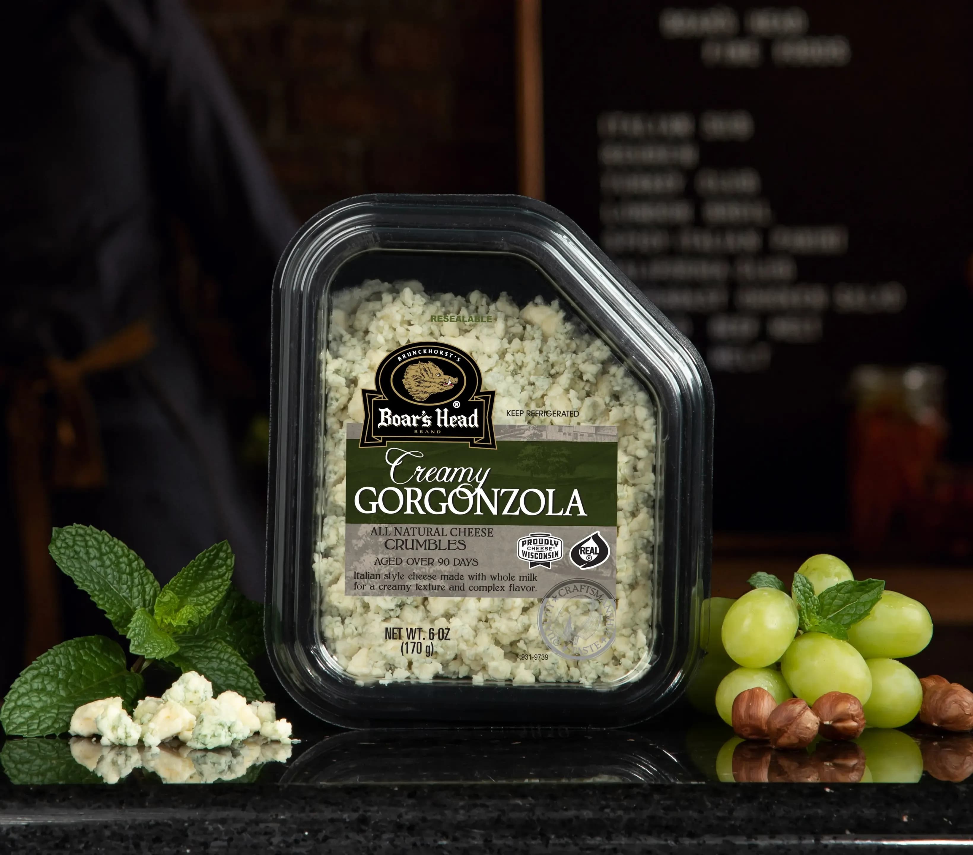 The Boar's Head® Creamy Gorgonzola Cheese Crumbles sits on a counter top, surrounded by grapes, nuts, greenery, and crumbled product, all in front of blurred deli scene in the background.