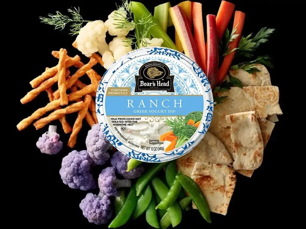 The Boar's Head® Ranch Greek Yogurt Dip sits on a counter top, surrounded by pretzels, cauliflower, green beans, pita, slices of peppers, celery sticks, and other colorful vegetables for dipping.