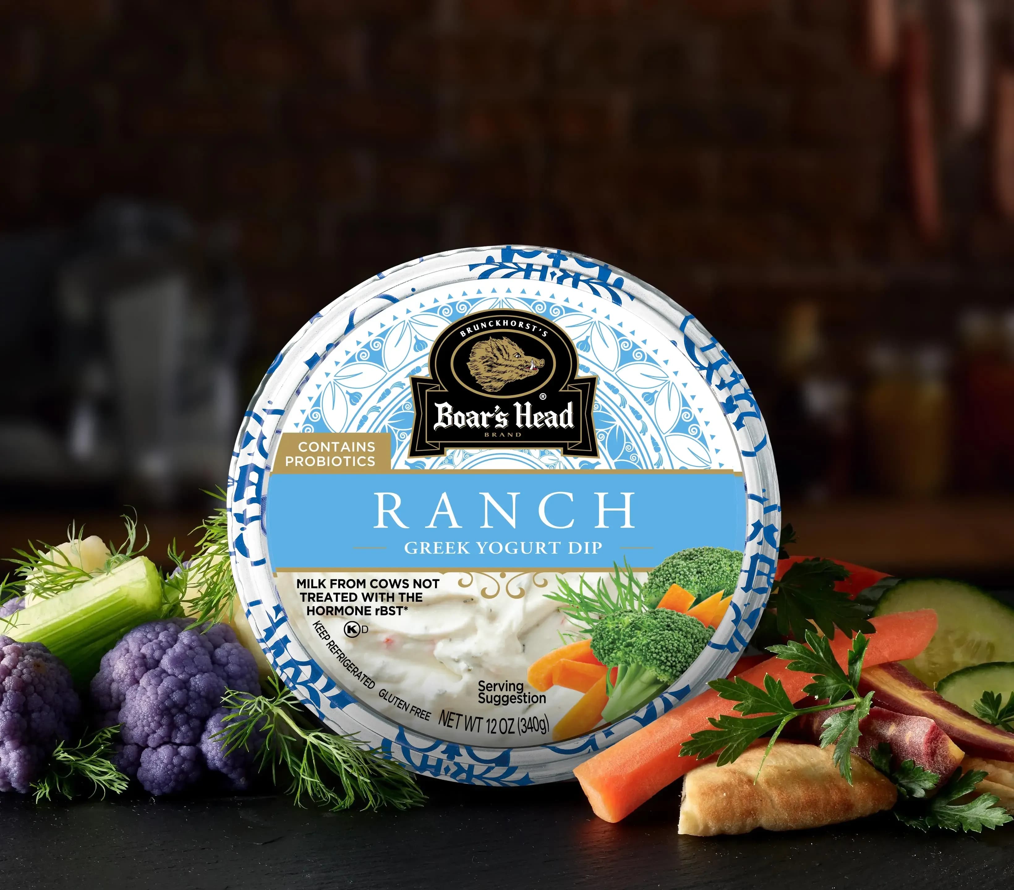 The Boar's Head® Ranch Greek Yogurt Dip sits on a counter top, surrounded by cauliflower, green beans, pita, slices of peppers, celery sticks, and other colorful vegetables for dipping, all in front of blurred deli background.