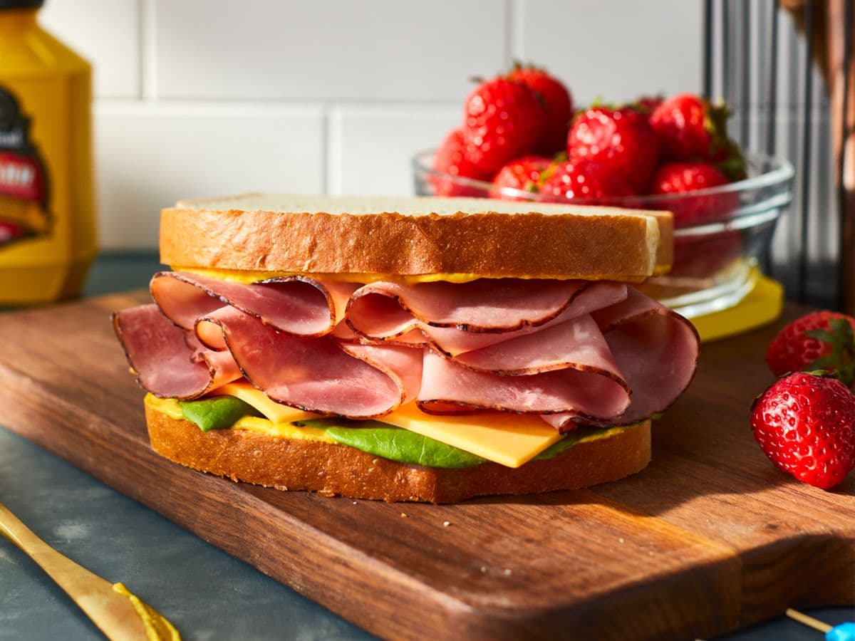 A sandwich with layers of sliced ham, cheese, and lettuce, served on two slices of bread, alongside fresh strawberries and mustard in the background.