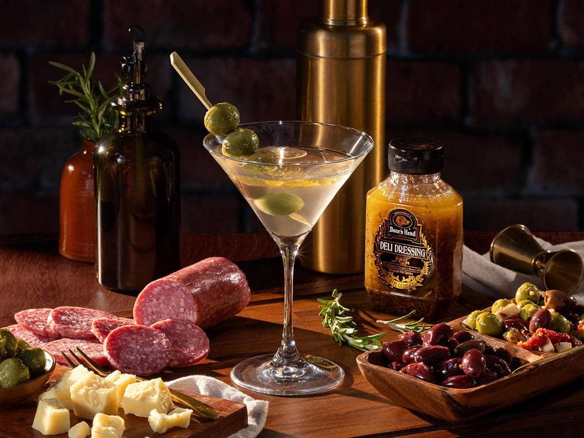 A martini made from brine and olives from the Boar's Head® collection sits on a table surrounded by items like sliced cheeses, olives, sliced charcuterie, Deli dressing, a martini shaker, and a plant in a vase.