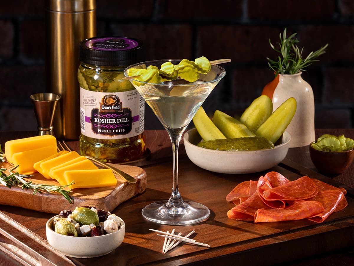 A martini made from brine and olives from the Boar's Head® collection sits on a table surrounded by items like sliced cheese, olives, sliced charcuterie, a bowl of halved pickled, a container of Boar's Head® pickles, and a plant in a vase.