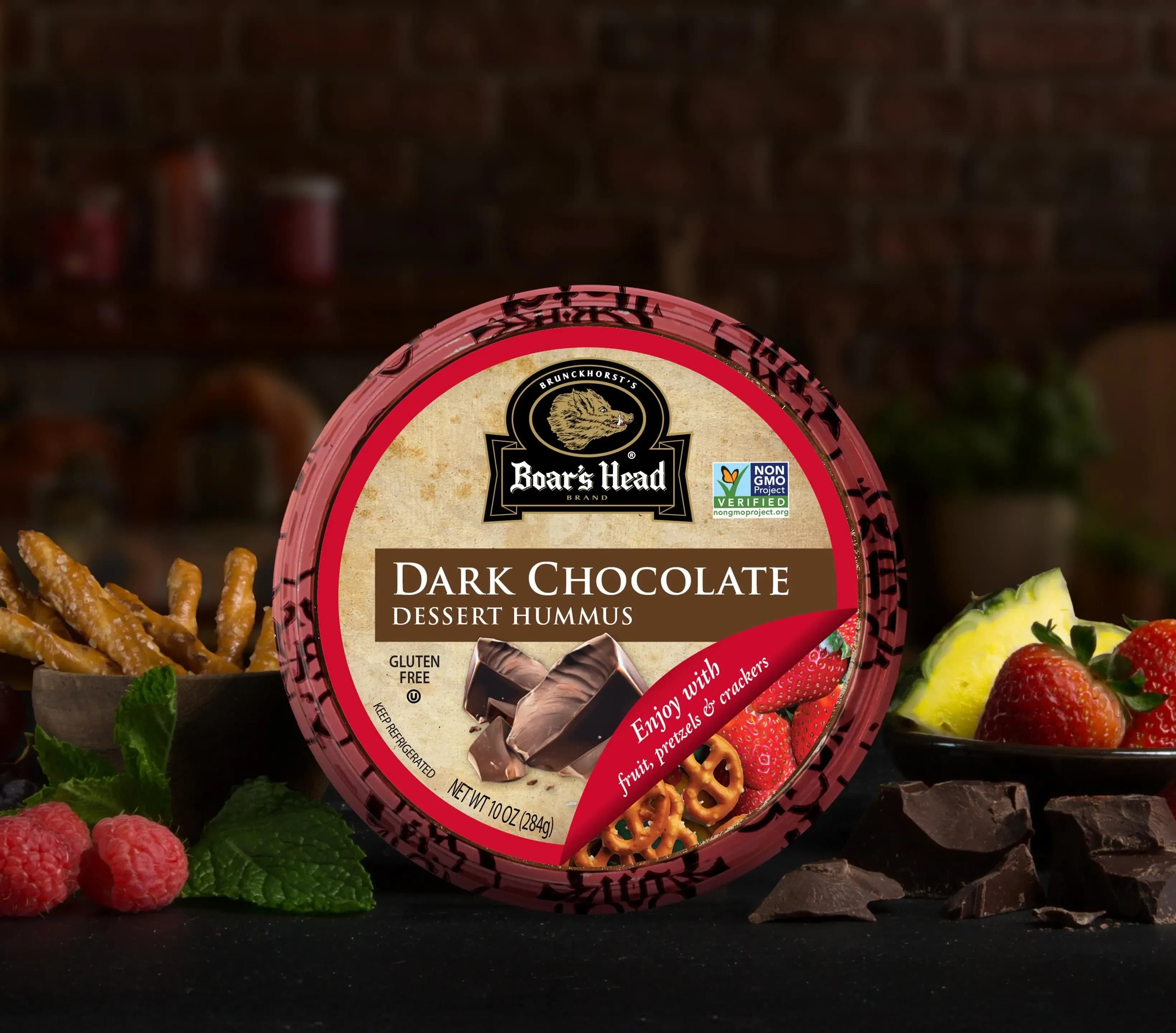 The Boar's Head® Dark Chocolate Hummus sits on a counter top, surrounded by dark chocolate pieces, raspberries, strawberries, pineapple slices, and a bowl of pretzel sticks.